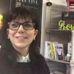 Woman wearing glasses is smiling in a room with books, decorative items, and a chalkboard wall.