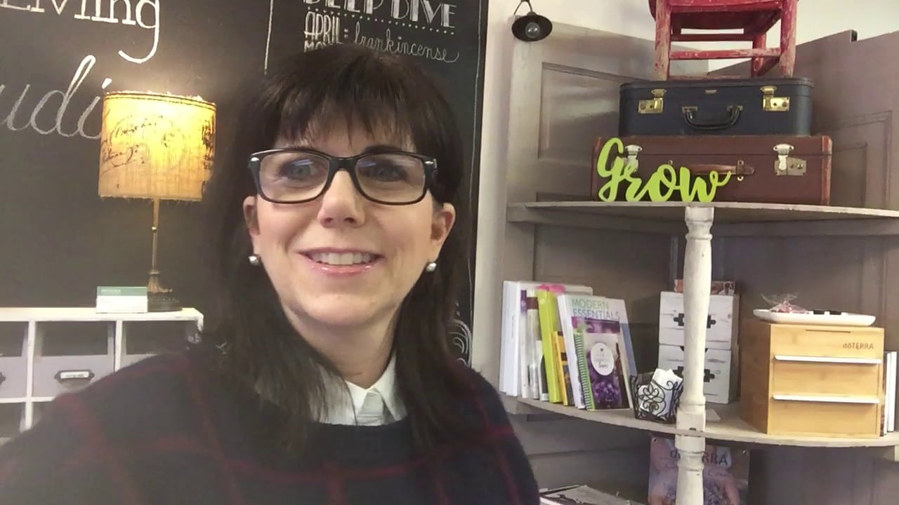 Woman wearing glasses is smiling in a room with books, decorative items, and a chalkboard wall.