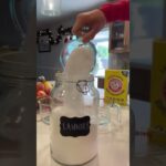 A person pours white powder from a measuring cup into a jar labeled "Laundry" next to a box of Arm & Hammer Super Washing Soda in a kitchen setting.