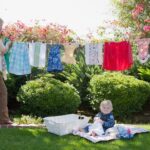 Person hanging clothes on a clothesline in a garden, with a baby sitting on a blanket nearby.