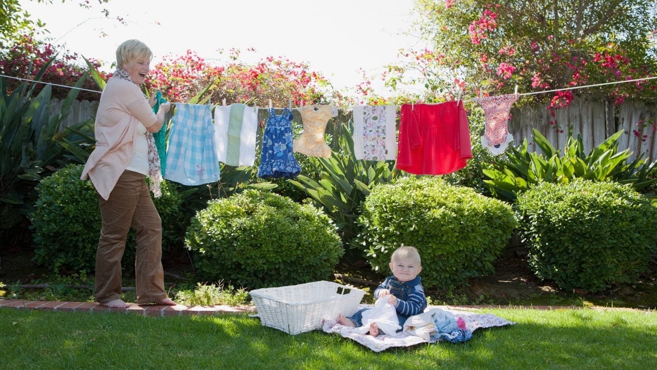 Person hanging clothes on a clothesline in a garden, with a baby sitting on a blanket nearby.